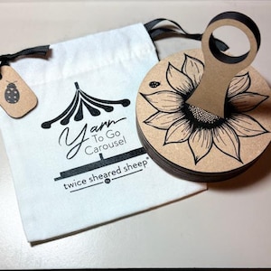 May include: A wooden yarn carousel with a sunflower design and a handle, next to a white bag with the text "Yarn To Go Carousel" and "twice sheared sheep". A small tag with a ladybug is attached to the bag.