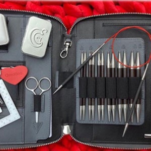 May include: A black zipped case containing knitting equipment, including needles, scissors, a tape measure, and a gauge tool. The case is open, displaying the contents against a red, textured backdrop. The needles are silver and black.