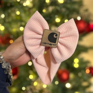 May include: A light pink baby headband with a large bow. The bow features a textured fabric and a brown leather patch in the center with the Carhartt logo in black and white. The headband is held up against a blurred background of Christmas lights.