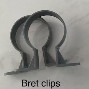 May include: Two grey Bret clips, designed to secure pipes or other cylindrical objects. Each clip has a semi-circular shape with a flat base and a mounting bracket. The text "Bret clips" is displayed at the bottom.