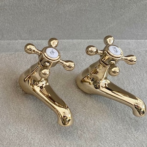 May include: Two polished gold-colored faucets with cross-shaped handles. Each faucet has a white circular center with the words "Hot" and "Cold" in black script. The faucets are mounted on a light gray surface.