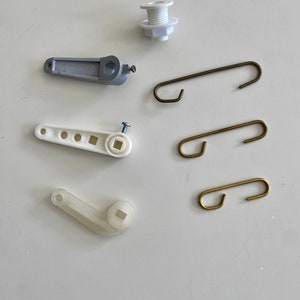 May include: A collection of plumbing hardware components, including white and grey plastic levers, a white threaded fitting, and several metal hooks in various sizes and colours. The parts are arranged on a white surface.