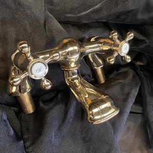 May include: A polished chrome bathroom faucet with cross-shaped handles. The faucet has two spouts and features white porcelain accents with the letters "Hot" and "Cold". The fixture is set against a dark, textured background.