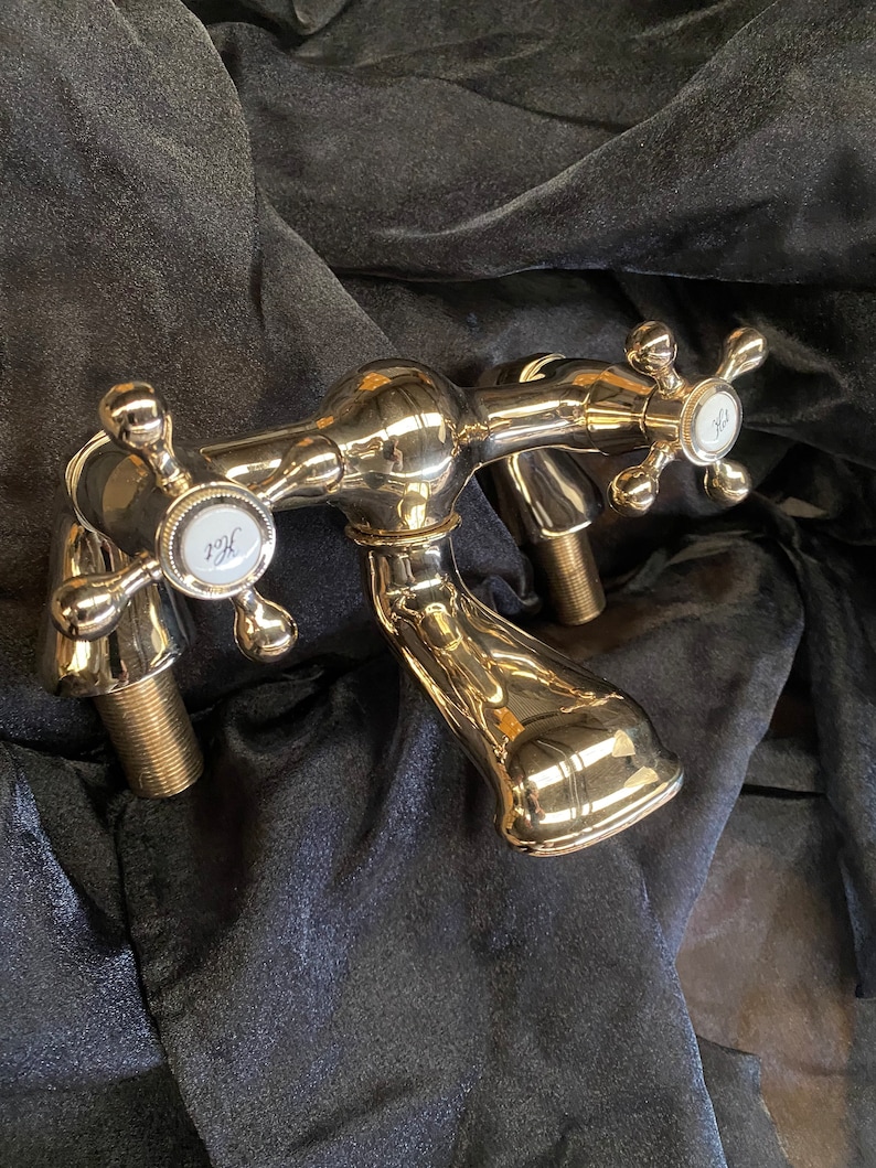 May include: A polished chrome bathroom faucet with cross-shaped handles. The faucet has two spouts and features white porcelain accents with the letters "Hot" and "Cold". The fixture is set against a dark, textured background.
