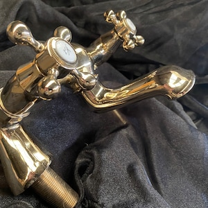 May include: A polished brass faucet with cross-shaped handles and a curved spout. The faucet has a shiny, reflective surface and a threaded base for installation. The handles have white accents. The overall design is vintage.