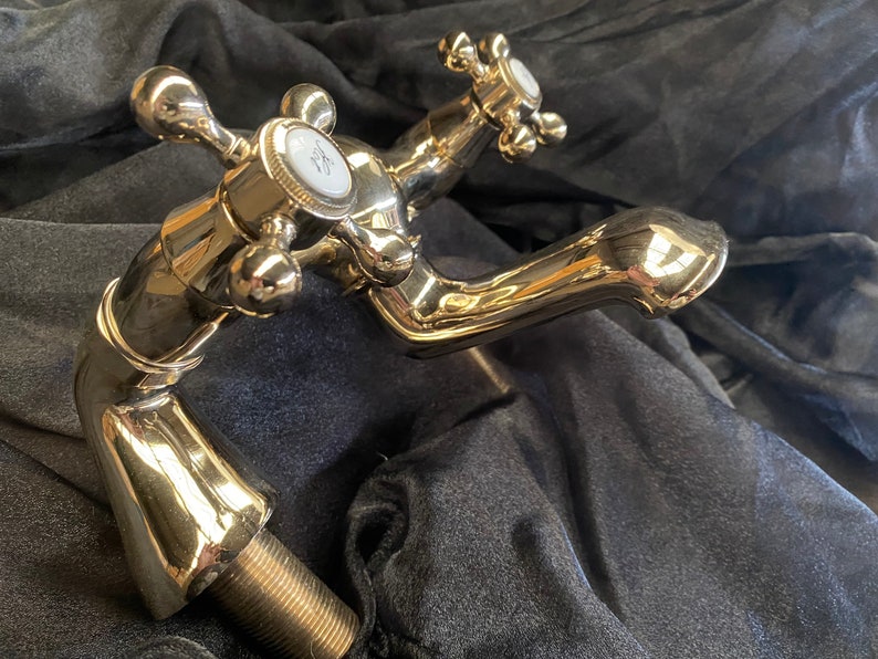 May include: A polished brass faucet with cross-shaped handles and a curved spout. The faucet has a shiny, reflective surface and a threaded base for installation. The handles have white accents. The overall design is vintage.
