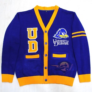May include: A blue and gold University of Delaware cardigan sweater. The sweater features the letters "UD" in gold, the university logo, and the words "UNIVERSITY OF DELAWARE". The sweater has gold trim and two gold stripes on the left sleeve.