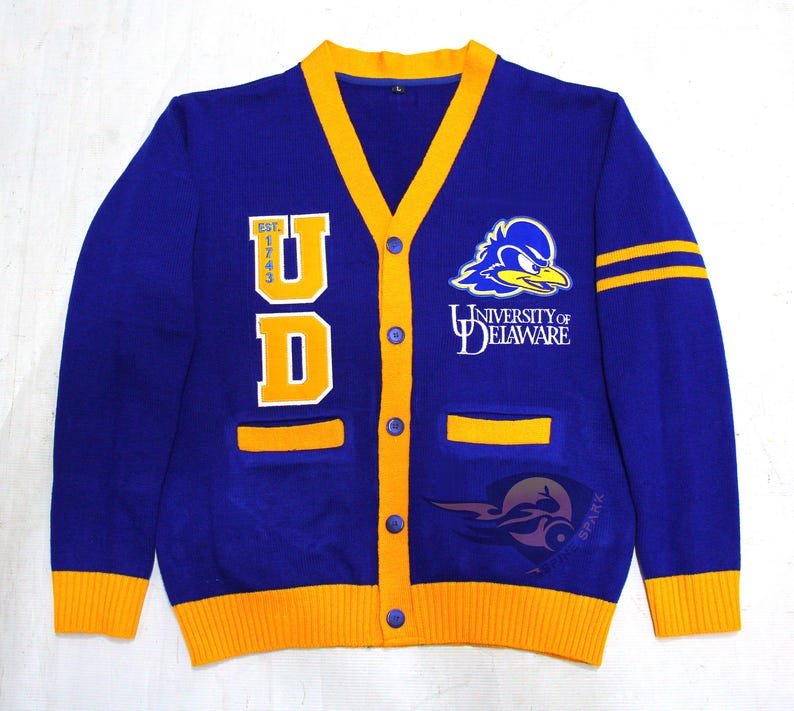 May include: A blue and gold University of Delaware cardigan sweater. The sweater features the letters "UD" in gold, the university logo, and the words "UNIVERSITY OF DELAWARE". The sweater has gold trim and two gold stripes on the left sleeve.