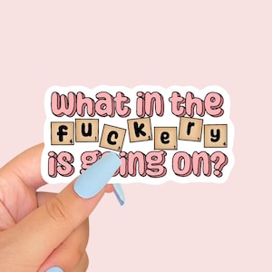 Peut inclure: Un autocollant rose et blanc avec le texte "What in the f*ckery is going on?" Le texte "f*ckery" est dans des cases individuelles, comme une partie de Scrabble.