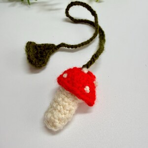 May include: A crocheted mushroom ornament. The mushroom has a red cap with white spots, a cream-colored stem, and a green stem and leaf. A charming, handmade decorative item.