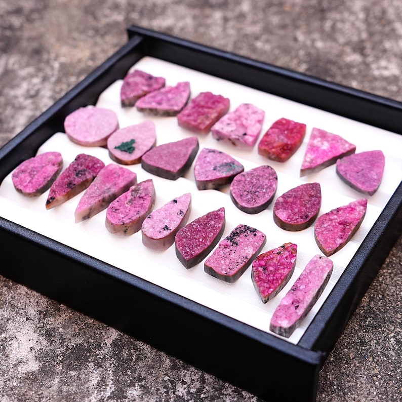 May include: A collection of polished gemstones in shades of pink and magenta, displayed in a black tray with a white interior. The stones vary in shape, including teardrops, ovals, and irregular forms. Some stones have dark speckles.