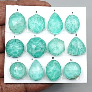 Puede incluir: Una colección de doce piedras preciosas de amazonita pulidas en varias formas, incluyendo ovaladas, hexagonales, de lágrima y redondas. Las piedras son de color turquesa claro con vetas sutiles, exhibidas sobre una superficie blanca.