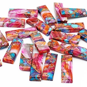 May include: A collection of rectangular gemstone beads in various colors. The beads feature a mix of pink, red, orange, blue, and gold hues. The gemstones are arranged on a white surface.