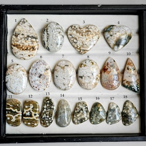 May include: A collection of polished gemstones in various shapes, including oval, teardrop, and heart. The stones display a mix of colors, such as white, brown, and green, with unique patterns and textures. The gemstones are arranged in a black display case.