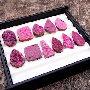 May include: A collection of polished, raw gemstones in various shapes and shades of pink and magenta, displayed in a black tray with a white interior. The gemstones include oval, rectangular, and teardrop shapes, with visible textures and patterns.