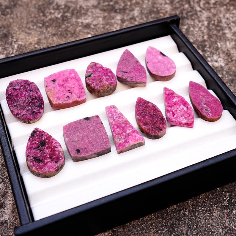 May include: A collection of polished, raw gemstones in various shapes and shades of pink and magenta, displayed in a black tray with a white interior. The gemstones include oval, rectangular, and teardrop shapes, with visible textures and patterns.