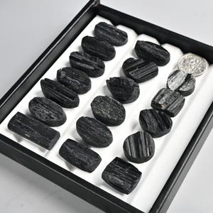 May include: A collection of polished black tourmaline stones displayed in a black tray with a white interior. The stones vary in shape, with some oval and others rectangular. A US dime is included for size reference.