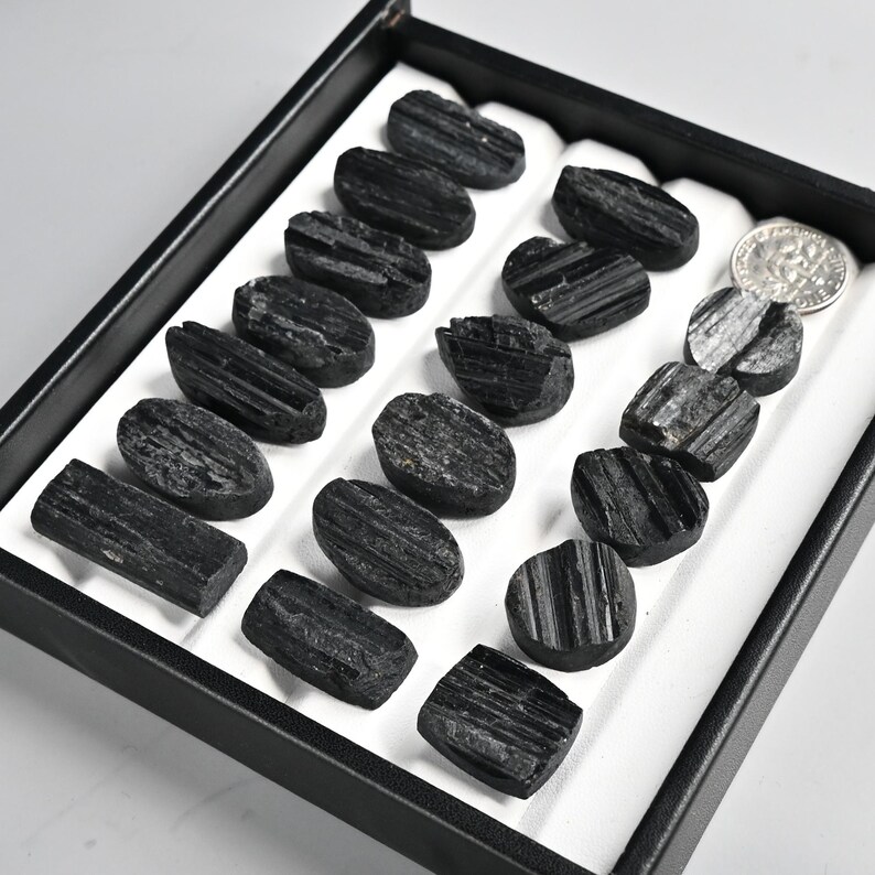 May include: A collection of polished black tourmaline stones displayed in a black tray with a white interior. The stones vary in shape, with some oval and others rectangular. A US dime is included for size reference.