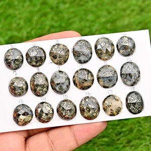 Könnte beinhalten: Eine Sammlung von achtzehn ovalen Pyrit-Edelsteinen, jede mit einem einzigartigen Muster aus metallischen Gold- und Silberflocken. Die Edelsteine werden auf einer weißen Karte vor grünem Hintergrund präsentiert.