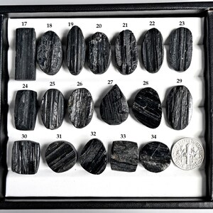 May include: A collection of black tourmaline gemstones in various shapes, including oval, rectangular, and teardrop. The gemstones are displayed in a black tray with a dime for size comparison. Each stone is numbered from 17 to 34.