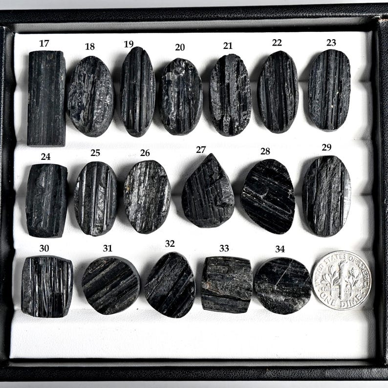 May include: A collection of black tourmaline gemstones in various shapes, including oval, rectangular, and teardrop. The gemstones are displayed in a black tray with a dime for size comparison. Each stone is numbered from 17 to 34.