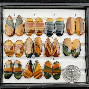May include: Eleven polished tiger's eye gemstones in assorted shapes, including oval and teardrop. The stones showcase golden brown, blue, and green colours. A quarter coin is included for scale. The gemstones are displayed in a black tray.