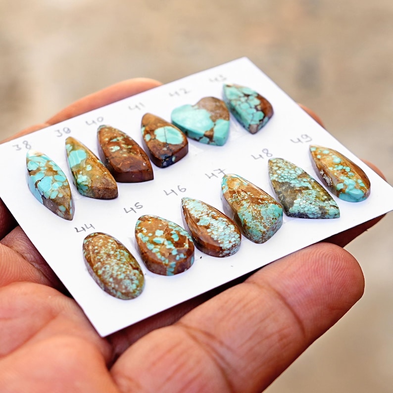 Puede incluir: Una colecci&oacute;n de doce cabujones de turquesa pulidos con patrones de matriz marr&oacute;n, dispuestos sobre una tarjeta blanca etiquetada con los n&uacute;meros del 38 al 49. Cada piedra presenta tonos azul-verdosos &uacute;nicos y vetas distintas.