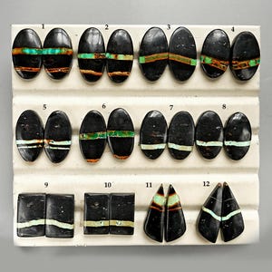 May include: A collection of twelve polished gemstone cabochons in various shapes. The stones are primarily black with bands of turquoise, green, and orange. The shapes include ovals, rectangles, and triangles, displayed on a white surface.