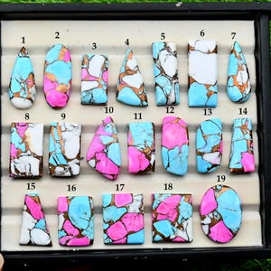 Puede incluir: Una colección de diecinueve cabujones de piedras preciosas turquesas, rosas y blancas en varias formas, incluyendo lágrima, ovalada y rectangular. Las piedras tienen una matriz de vetas color cobre. Los cabujones se muestran en una bandeja negra.