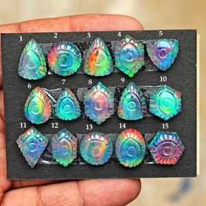 Puede incluir: Quince piedras preciosas talladas e iridiscentes dispuestas sobre una tarjeta negra, numeradas del 1 al 15. Cada piedra presenta un motivo central en forma de ojo con surcos radiantes, mostrando tonos cambiantes de azul, verde, naranja y rosa.