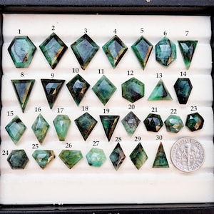 Puede incluir: Una colección de gemas de esmeralda en varias formas, incluyendo hexágonos, diamantes y triángulos. Las gemas son de color verde oscuro y están dispuestas en una bandeja. Se incluye una moneda para referencia de tamaño.