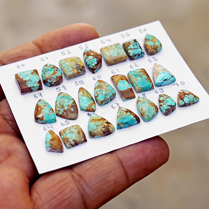 Puede incluir: Una colecci&oacute;n de veintiuna piedras preciosas de turquesa pulidas dispuestas sobre una tarjeta blanca. Cada piedra presenta un patr&oacute;n de matriz &uacute;nico de colores azul verdoso y marr&oacute;n. Las piedras est&aacute;n numeradas del 50 al 70 con tinta negra.