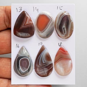 May include: Six polished cabochon gemstones arranged in two rows on a white card labeled with numbers 13 through 18. The stones feature varied banded patterns in shades of brown, orange, gray, and white, with pear and oval shapes.