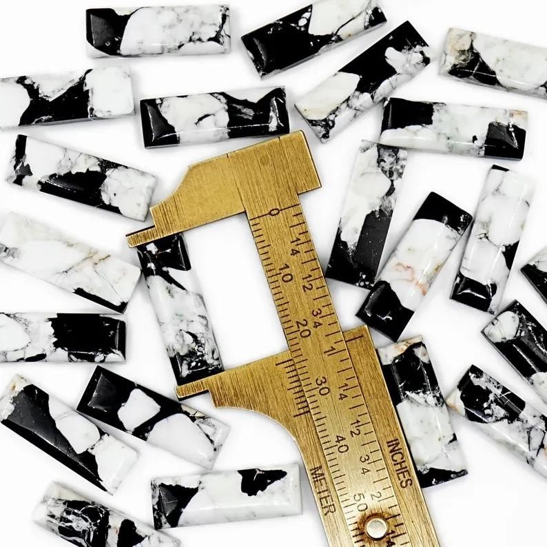 May include: A collection of rectangular, polished stones with a black and white marble pattern. A brass ruler with inch and centimeter markings is placed among the stones, indicating size. The stones are approximately 1 inch in length.