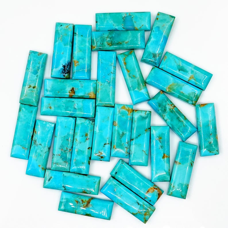 May include: A collection of rectangular turquoise gemstones with natural brown veining. The stones are polished and have a vibrant blue-green color. The gemstones are arranged on a white surface, showcasing their unique patterns and textures.