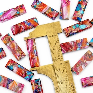 May include: A collection of rectangular gemstone beads in shades of red, pink, turquoise, and gold. A brass ruler with inch measurements is also present, indicating the size of the beads. The beads are scattered on a white surface.