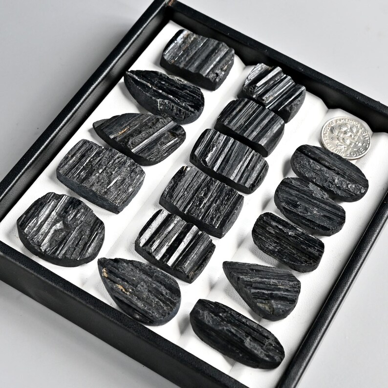 May include: A collection of polished black tourmaline stones, each with visible striations, arranged in a black tray with a white interior. A silver coin is also present. The stones vary in shape, with some rectangular and others teardrop-shaped.