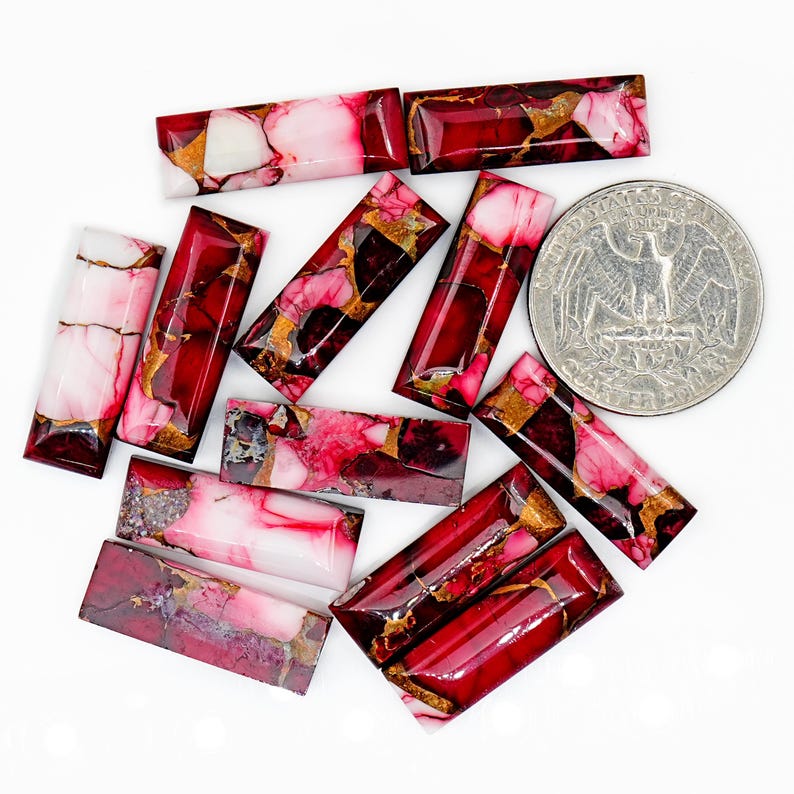 May include: A collection of rectangular gemstones with a mix of red, pink, white, and gold colors. The gemstones are arranged on a white surface, with a quarter coin for size comparison. The gemstones have a polished, glossy finish.