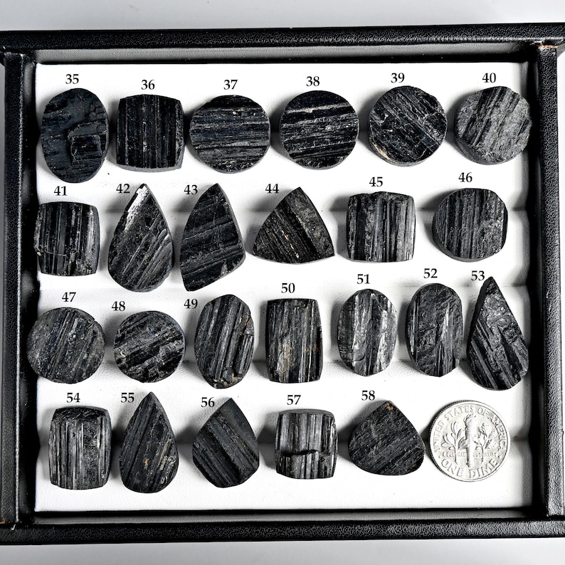 May include: A collection of polished black tourmaline gemstones in various shapes, including round, oval, and teardrop. Each stone is numbered and displayed in a black tray, with a dime for size comparison. The stones have a striped texture.