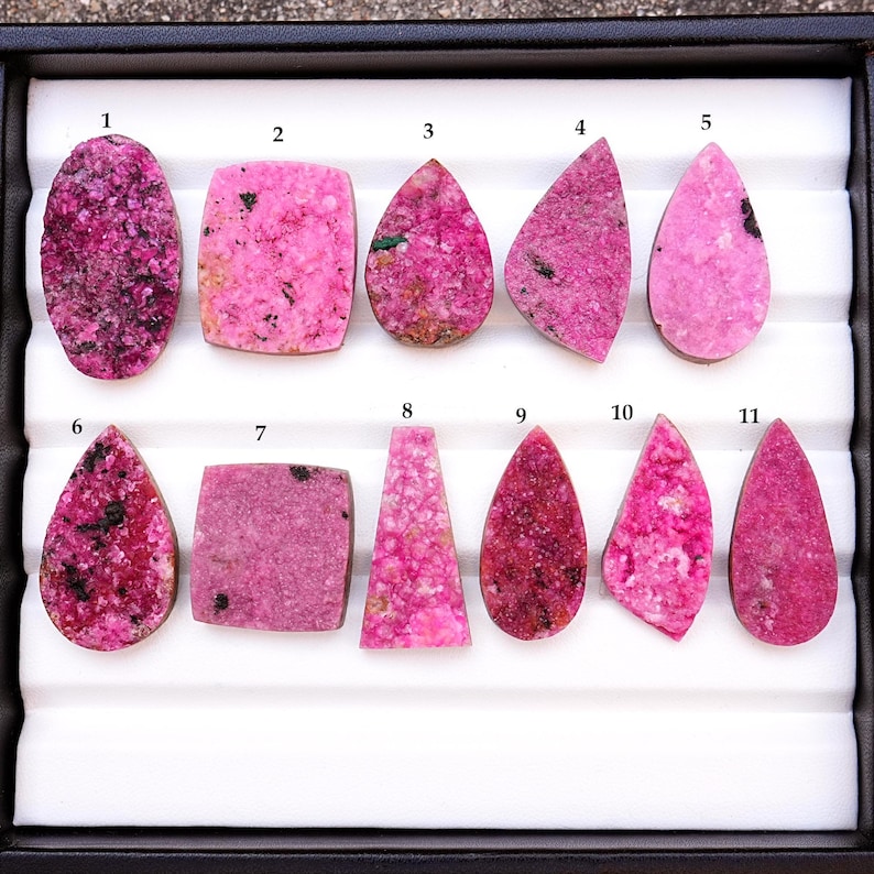 May include: A collection of eleven pink rhodonite gemstones in various shapes, including oval, rectangular, and teardrop. The stones have dark inclusions and are displayed in a black tray with a white interior.