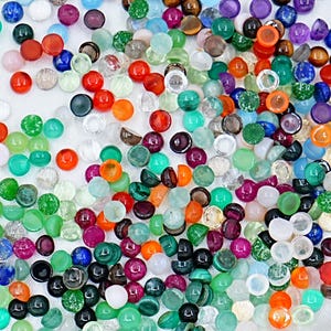 May include: A vibrant assortment of polished gemstones in a variety of colours and shapes. The gemstones include red, orange, green, blue, purple, and clear stones, with a shiny finish. The collection features round, oval, and irregular shapes, suitable for jewellery or craft projects.