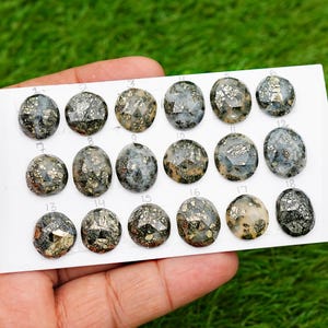 Op de afbeelding: Een verzameling van achttien ovale pyriet edelstenen, elk met een gefacetteerd oppervlak. De stenen tonen een mix van metallic goud- en grijstinten, tegen een witte achtergrond. De edelstenen zijn gerangschikt op een witte kaart.