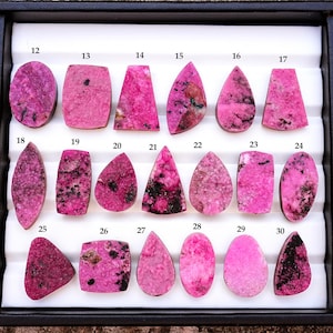 May include: A collection of polished rhodonite gemstones in various shapes, including ovals, rectangles, and teardrops. The stones display a vibrant pink hue with black inclusions, arranged in a black tray. Each stone is numbered.