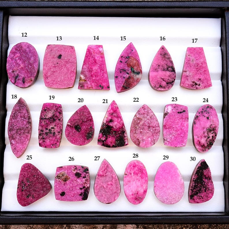 May include: A collection of polished rhodonite gemstones in various shapes, including ovals, rectangles, and teardrops. The stones display a vibrant pink hue with black inclusions, arranged in a black tray. Each stone is numbered.