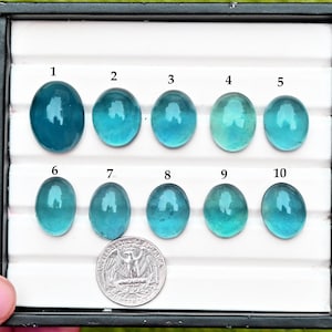 May include: Ten oval teal gemstones are displayed in a tray. A UK quarter coin is included for size comparison. The gemstones have a polished surface and vary in colour and clarity. The gemstones are measured in centimetres.