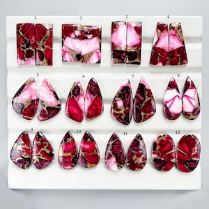 Puede incluir: Una colección de doce pares de cabujones de piedras preciosas pulidas de varias formas. Las piedras presentan una mezcla de colores rojo intenso, rosa, blanco y dorado. Los cabujones se exhiben sobre una superficie blanca.