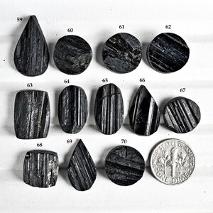 May include: A collection of black tourmaline gemstones in various shapes, including teardrop, oval, and round. The gemstones are polished and display natural striations. A US dime is included for size comparison.