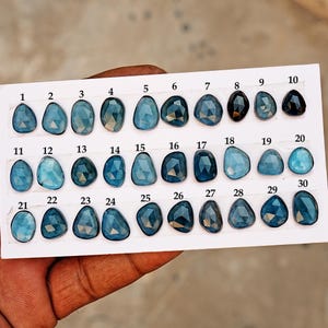 May include: A collection of thirty blue gemstones of various shapes and shades, arranged on a white card. Each stone is numbered from 1 to 30. The gemstones range in color from light teal to deep indigo.