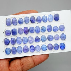 May include: An assortment of blue-violet gemstones in diverse shapes, such as oval, square, and teardrop. The gemstones are arranged in rows on a white card, displaying their different cuts and shades. The gemstones are held by a hand.