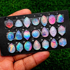 May include: A collection of twenty-one iridescent gemstone cabochons, each uniquely shaped and carved with intricate designs. The gemstones display a range of colors, including pink, blue, and green, and are arranged on a black surface.